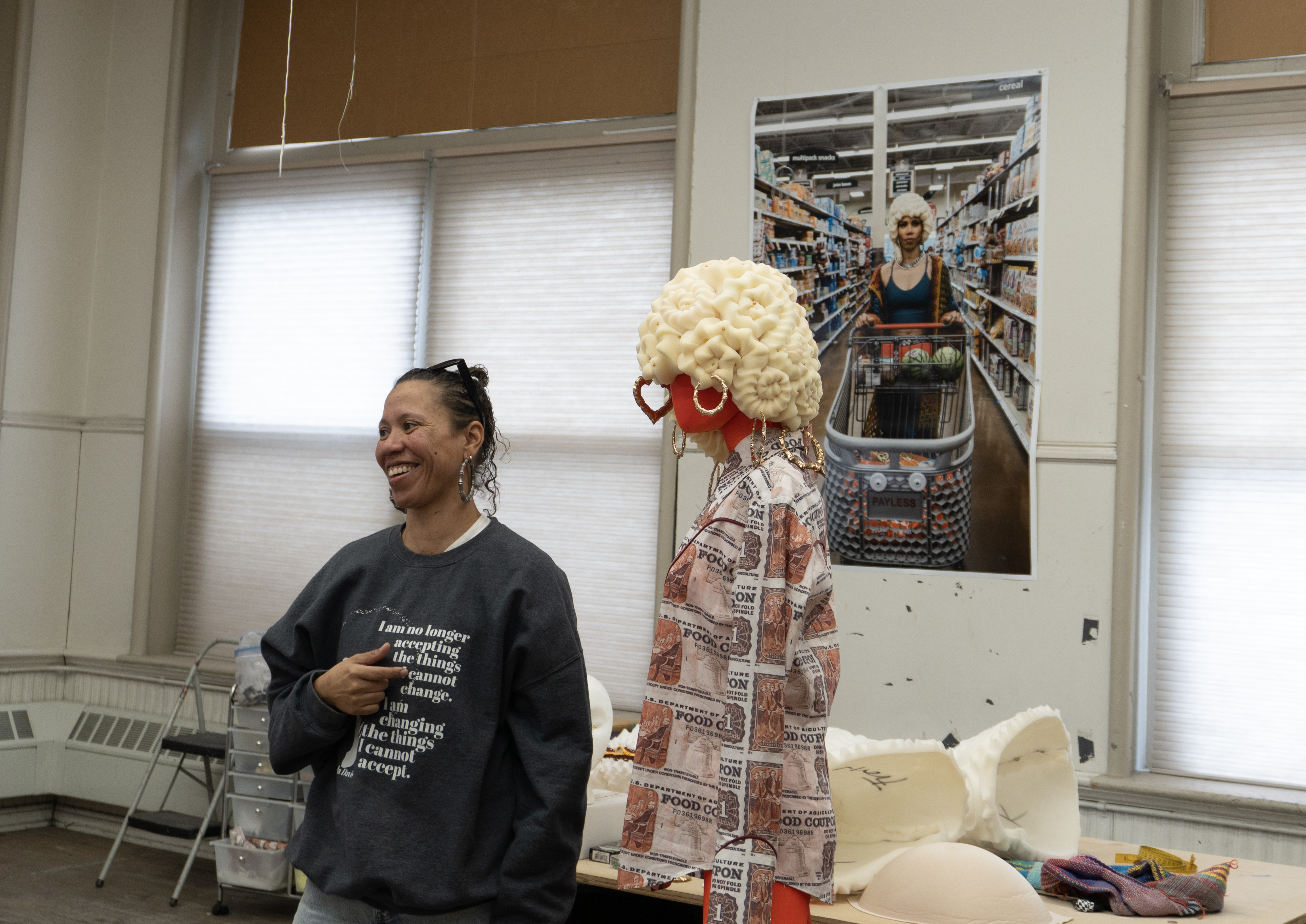 Desirae Brown smiles as she stands in her studio at ASLD next to a mannequin wearing two of her creations: a wig made from bed foam and bamboo hoop earrings, and a Western-style shirt made from fabric printed with a food stamp pattern.